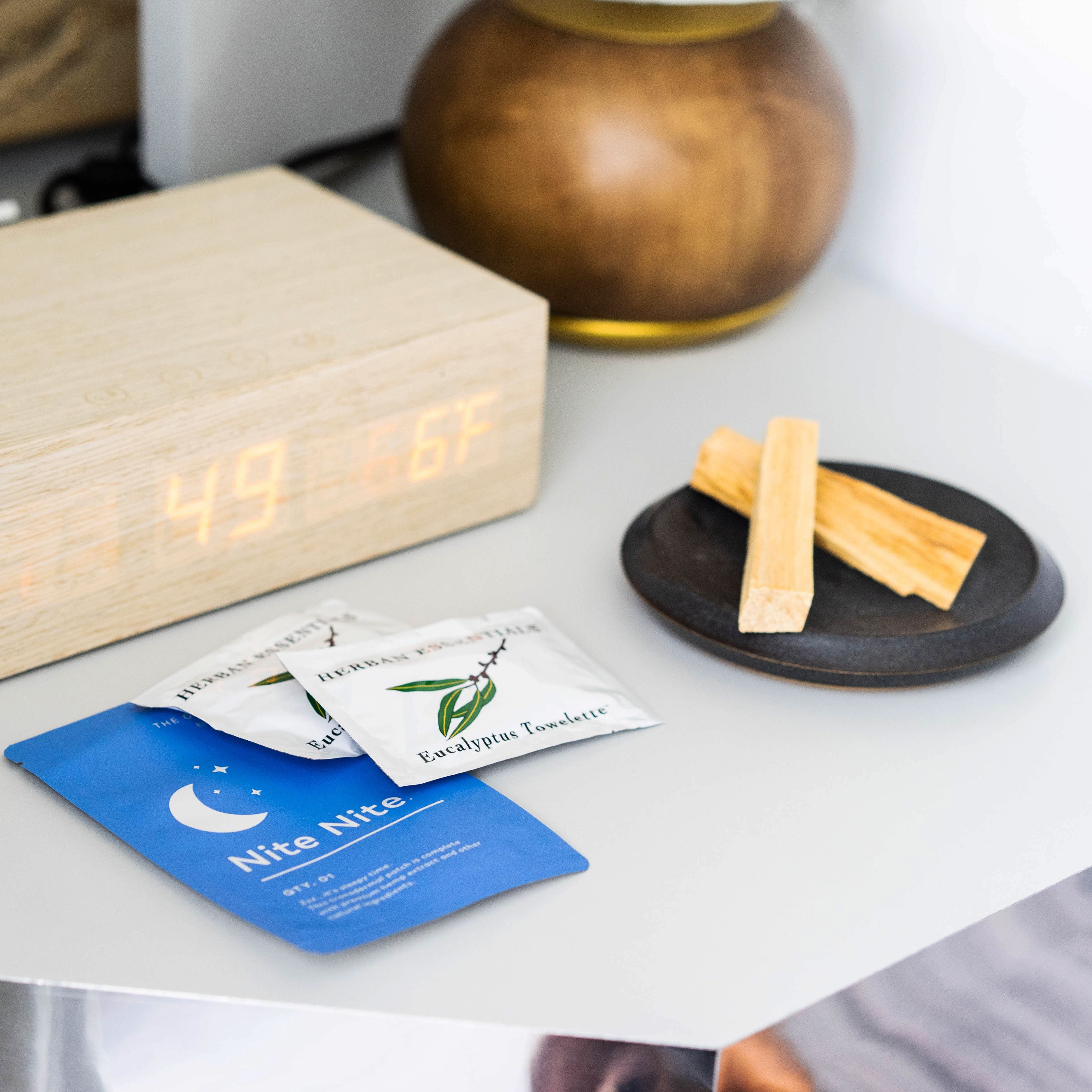 A wooden bedside table with a digital clock displaying 4:49, a packet of Eucalyptus tea, a blue card with a crescent moon, and two wooden sticks on a black coaster.