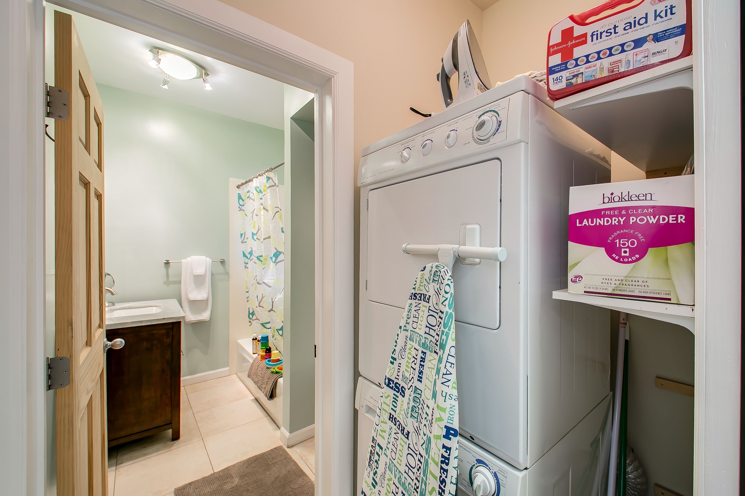 A laundry room with a front-loading washer and dryer, a box of Beko laundry powder, and a first aid kit on the shelf.