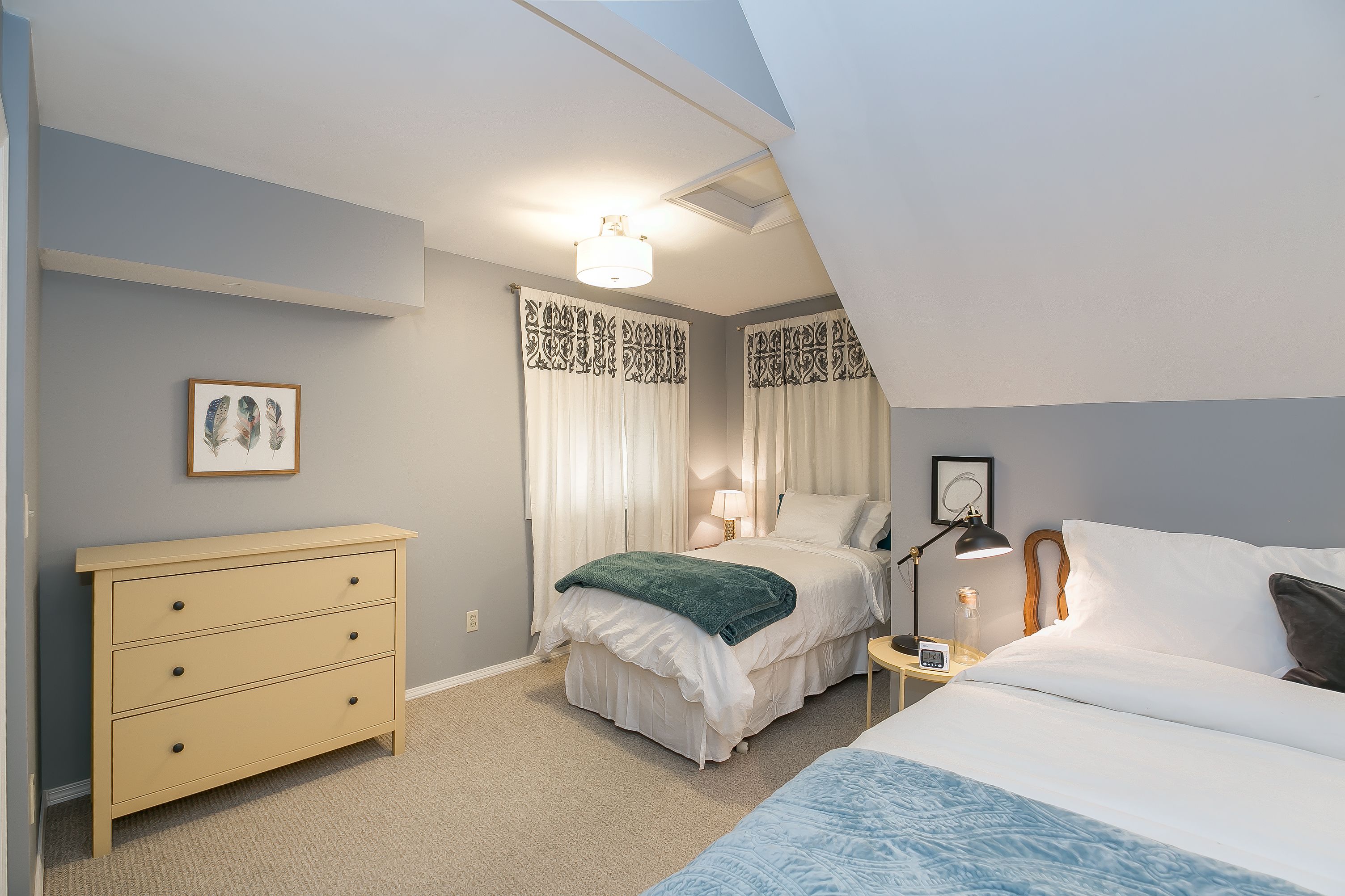 A bedroom with two beds, a dresser, a bedside table with a lamp, and a ceiling light.