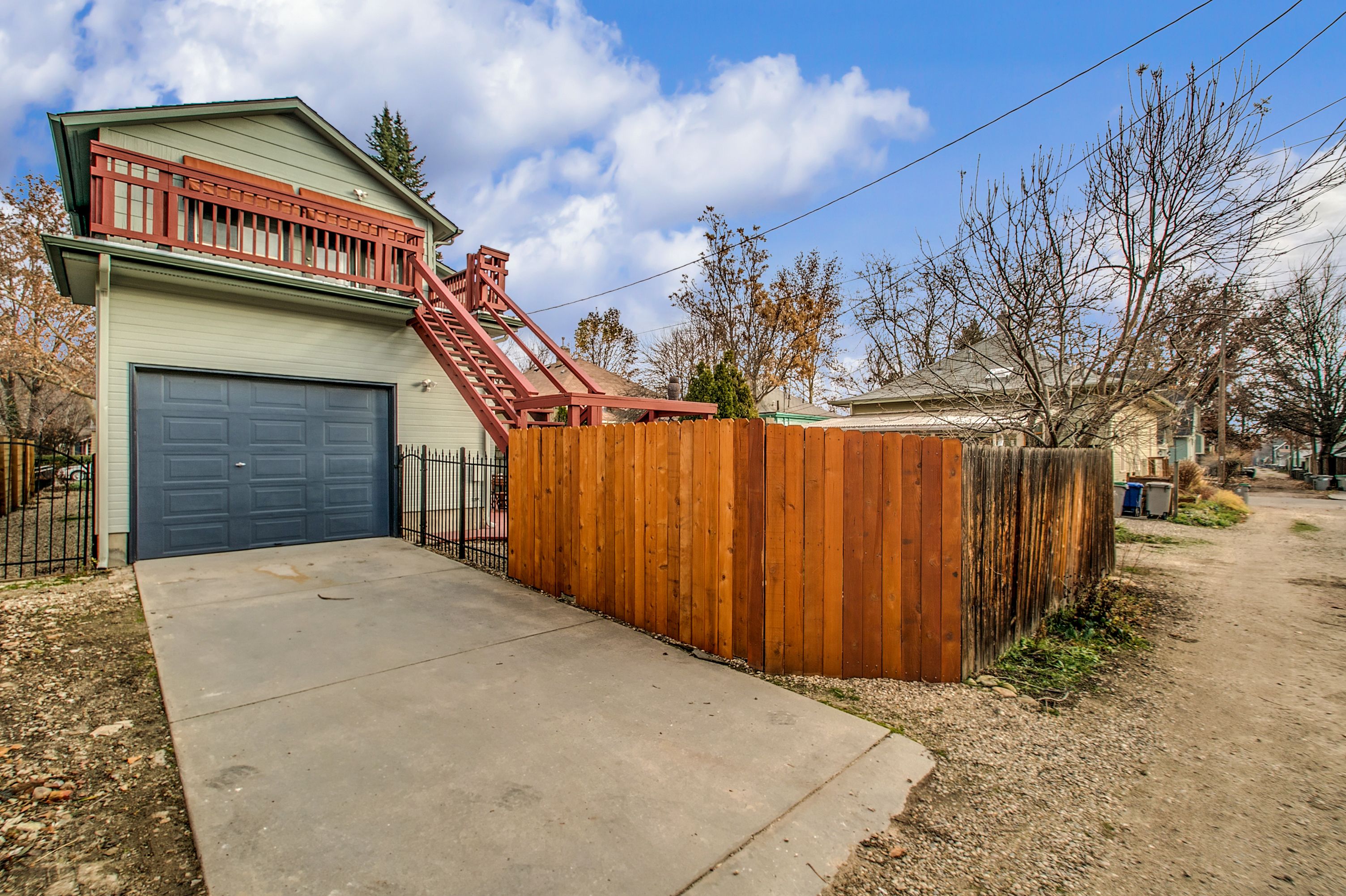 Un garage attenant à une maison, avec un escalier rouge menant à une terrasse, le tout entouré d’une clôture en bois et donnant sur une allée en béton.
