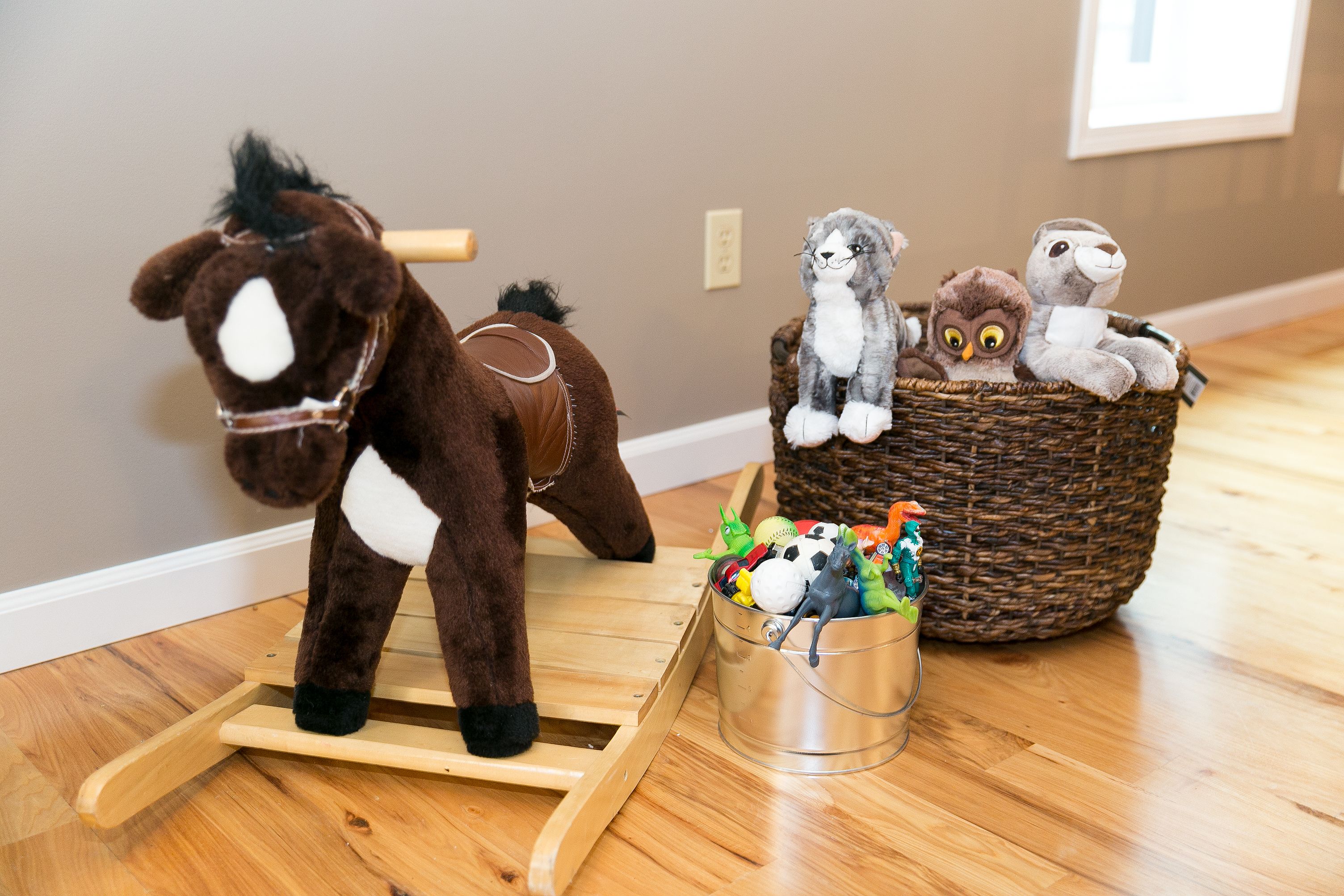 Un cheval à bascule en bois, un seau en métal rempli de jouets et un panier en osier contenant des peluches, le tout posé sur un plancher en bois.
