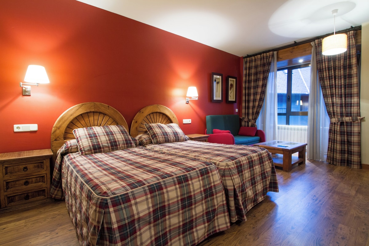 Une chambre avec un mur rouge, deux têtes de lit en bois, un lit avec une couette à carreaux, une table de chevet, un banc en bois, une chaise rouge et une fenêtre avec des rideaux.