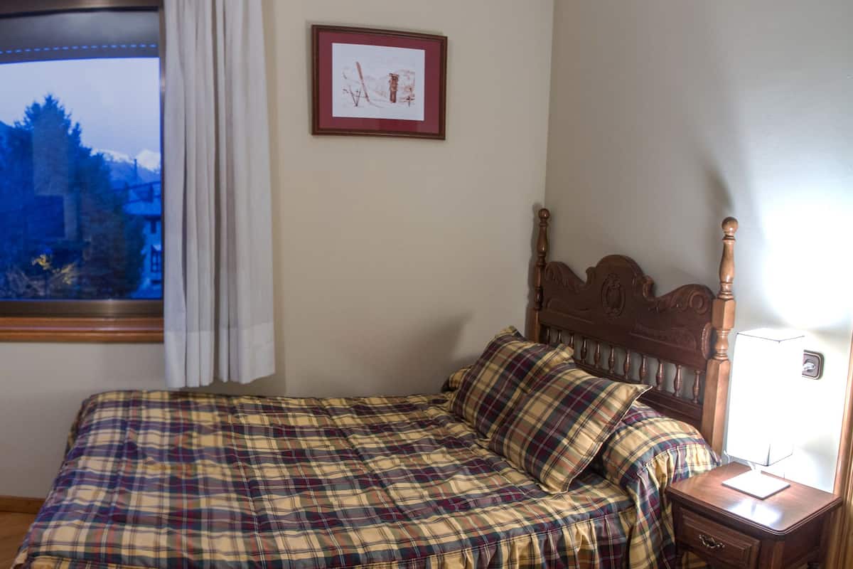Un dormitorio con cama, televisor sobre una mesa de madera, ventana con vista a las montañas y un cuadro en la pared.