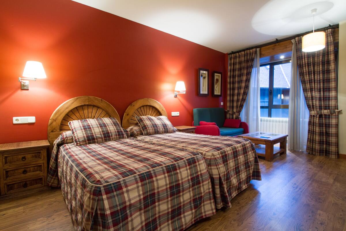 Un dormitorio con pared roja, dos cabeceros de madera, una cama con ropa de cama escocesa, una mesita de noche, un banco de madera, una silla roja y una ventana con cortinas.