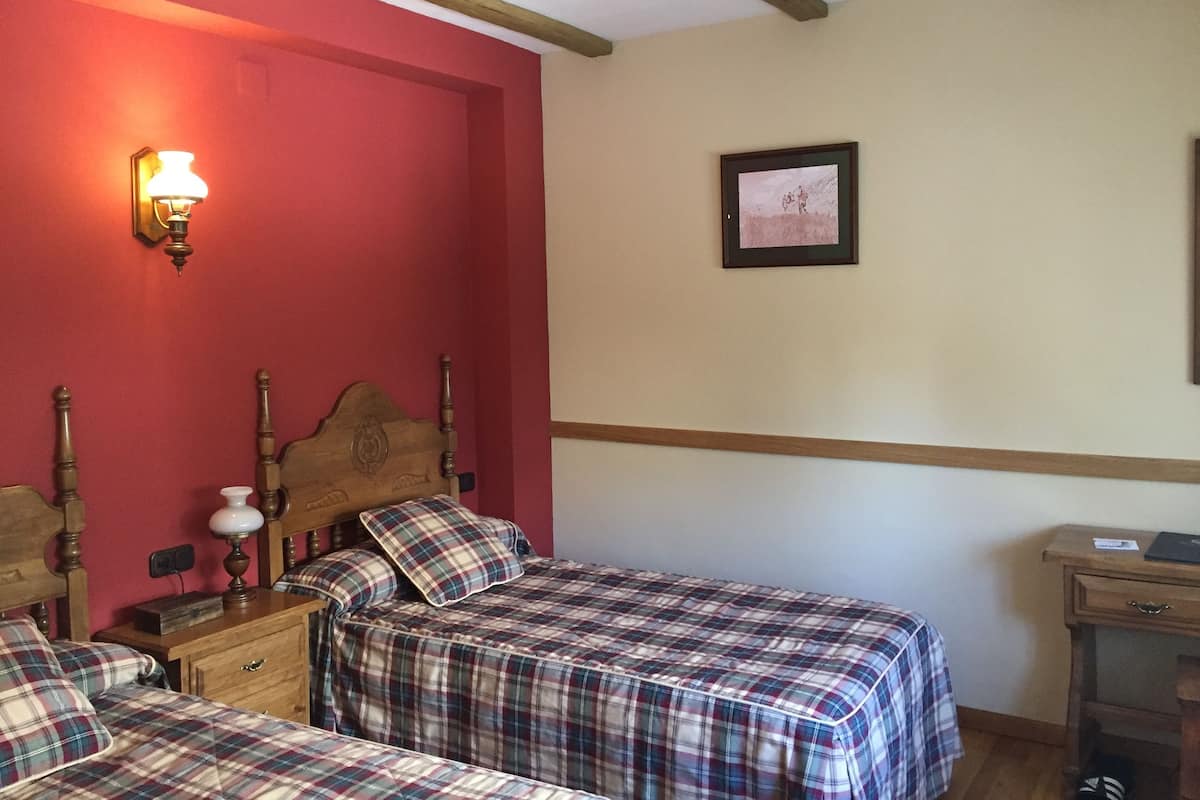 Una habitación con dos camas, un cabecero de madera, una mesita de noche con una lámpara, un cuadro enmarcado en la pared y una pequeña mesa de madera.