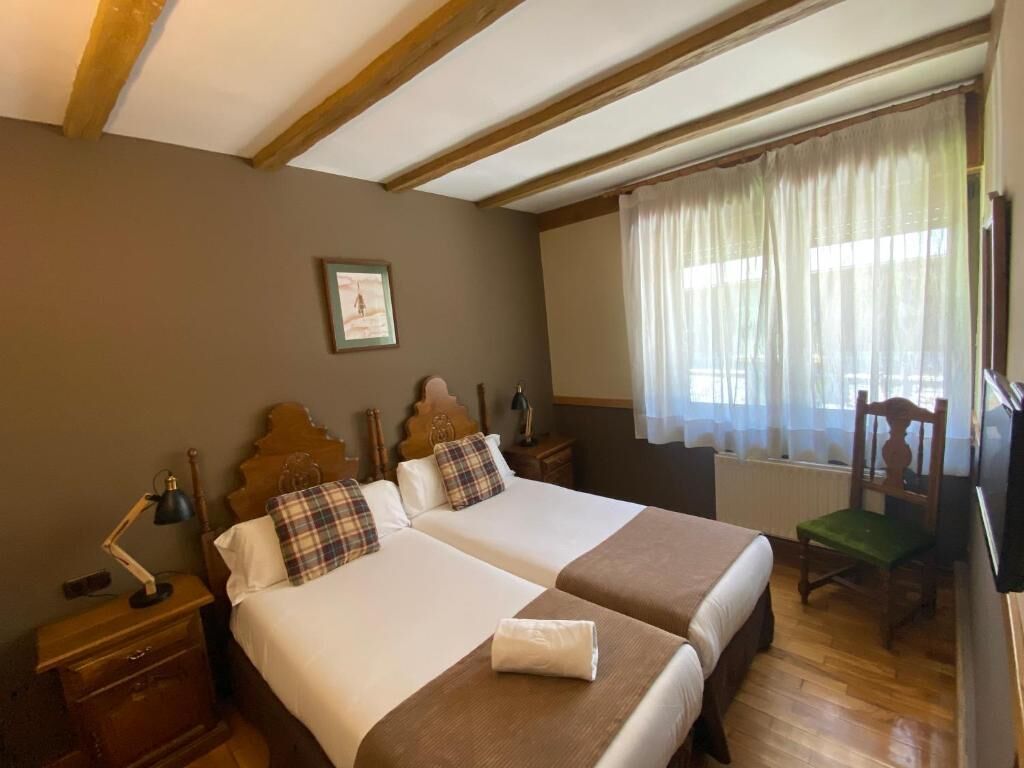 A double room with two beds, wooden headboards, a nightstand with a lamp, a chair, and a window with curtains.