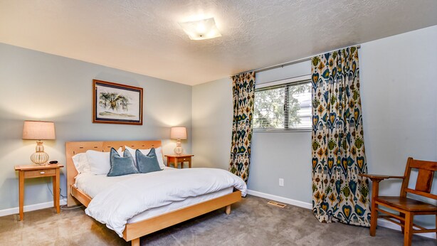 A bedroom with a bed, two bedside tables with lamps, a chair, a window with patterned curtains, and a wall-mounted picture. - The East End Retreat (Boise)