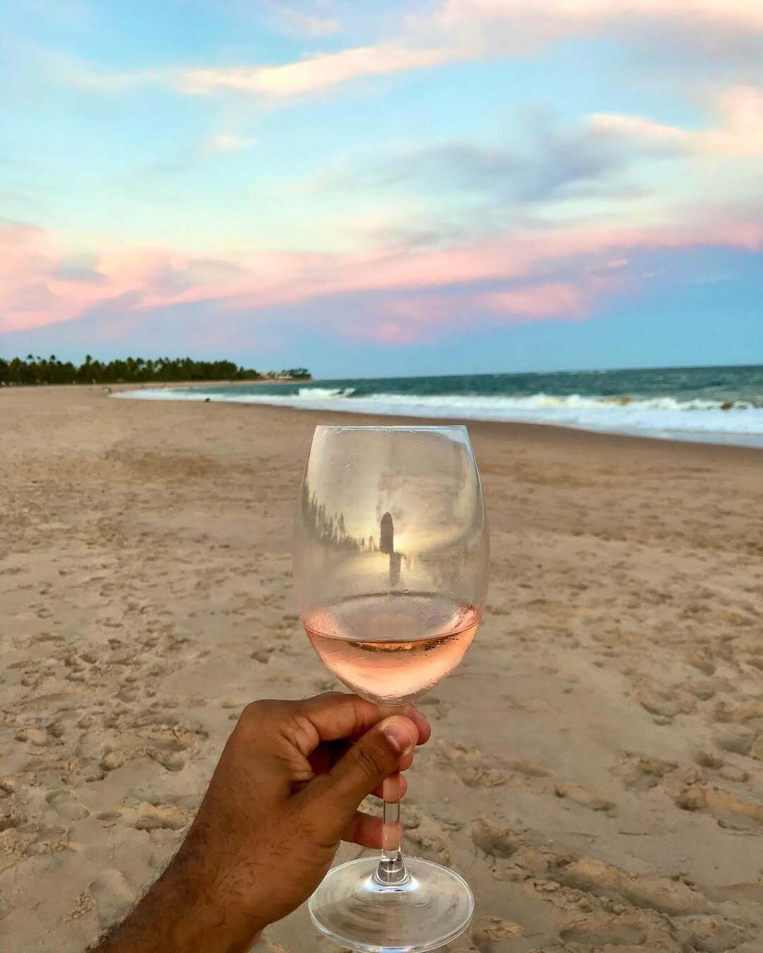 A beach scene with a clear glass of wine held in a hand.