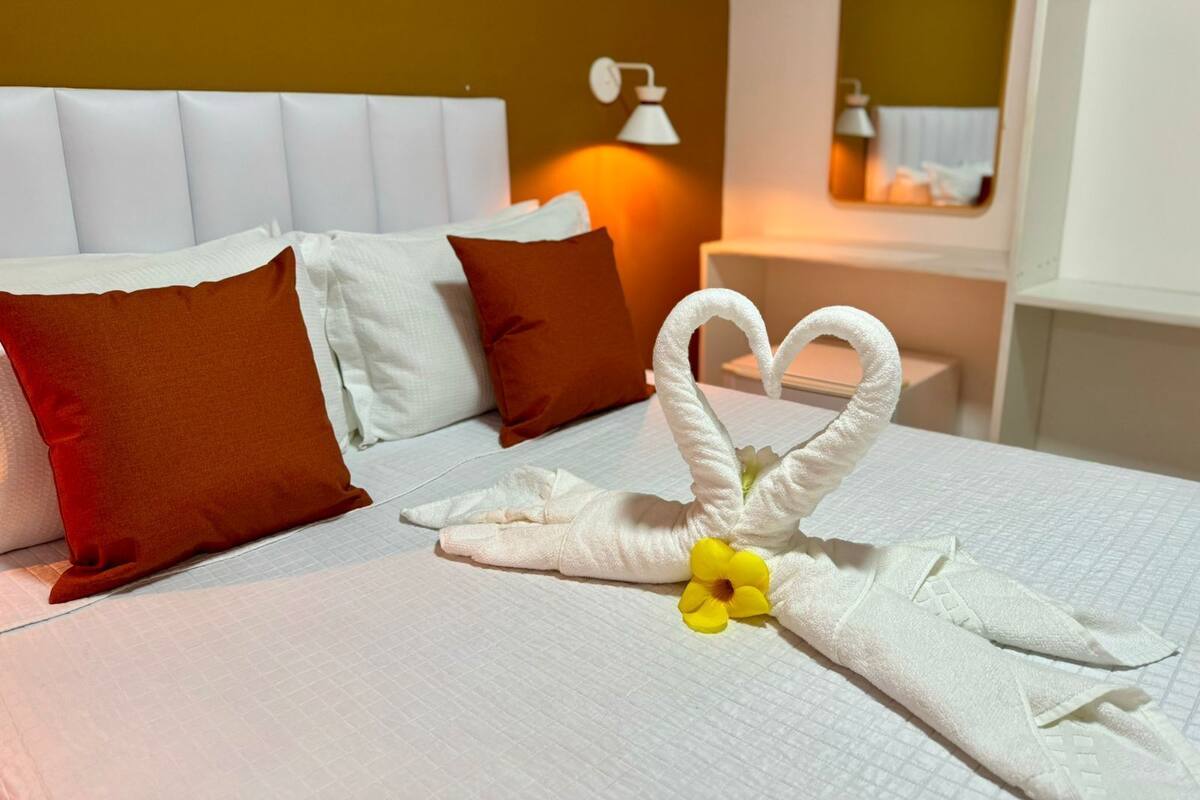 Een net opgemaakt bed met witte en bruine kussens, een handdoek in de vorm van een hart en een bedlampje.