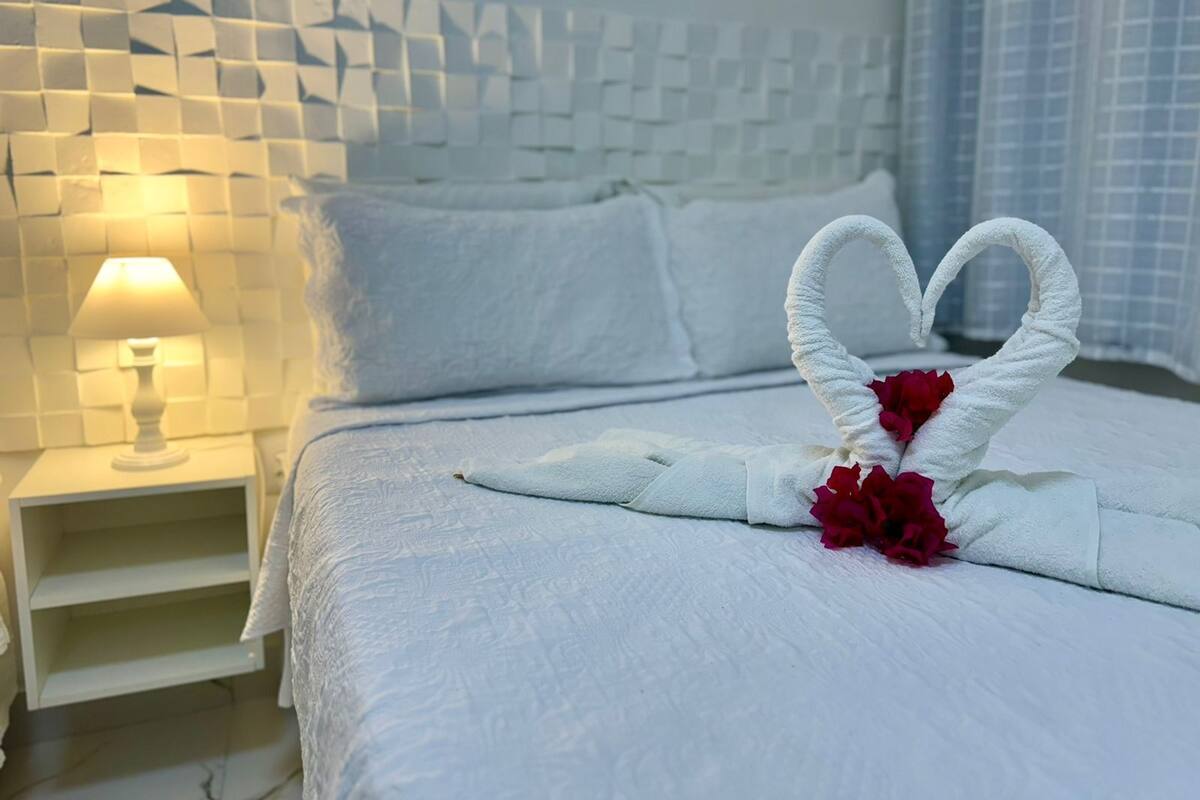 Een net opgemaakt bed met een handdoek in de vorm van een hart en rode bloemen, een bedlampje, en een muur met een geometrisch patroon.