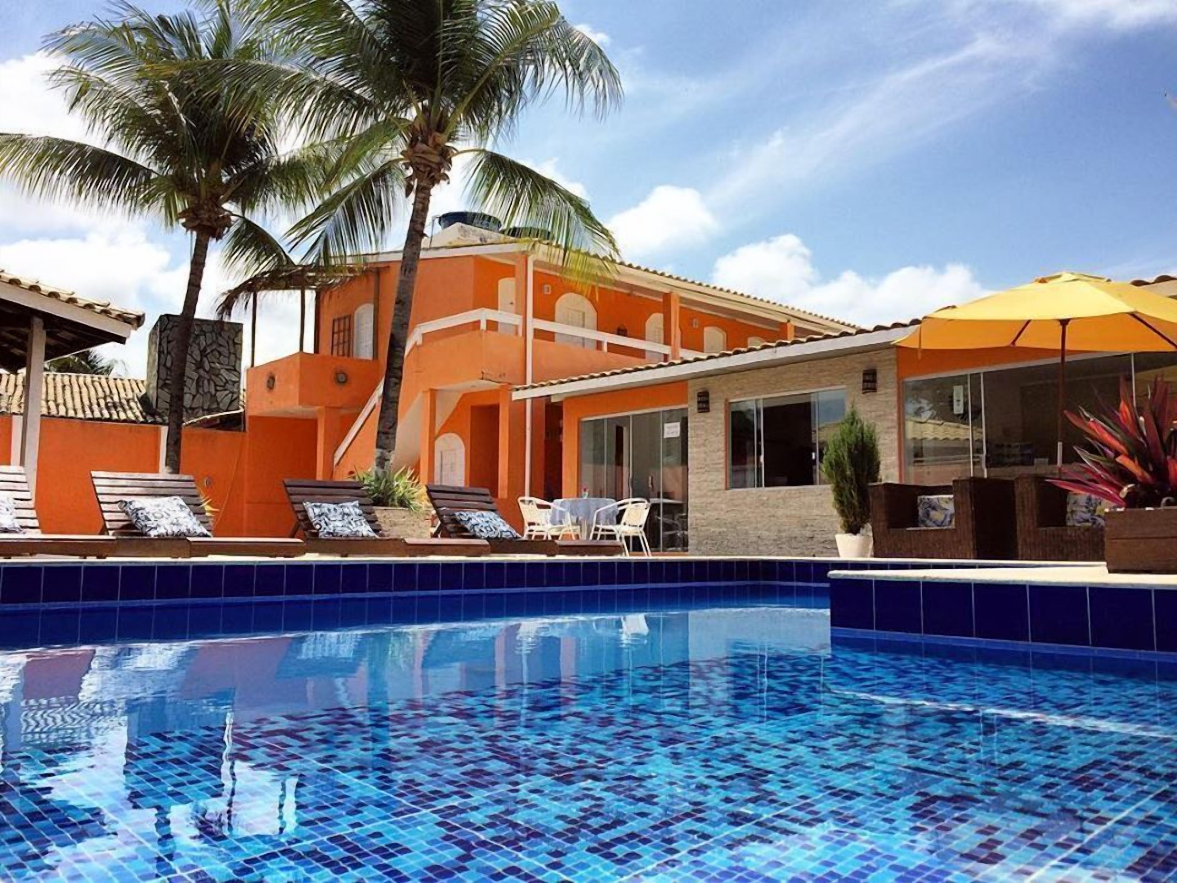 A swimming pool with blue mosaic tiles, a palm tree, and a building with orange walls and a tiled roof.