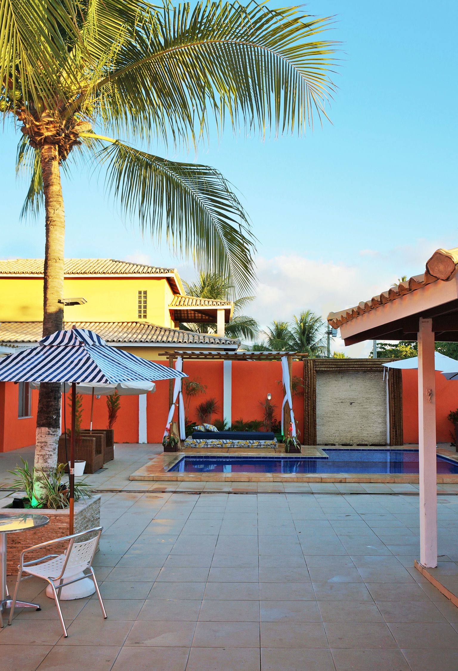 Un área de piscina con suelo de baldosas, una palmera, un paraguas a rayas y un edificio de fachada amarilla.