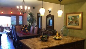 A kitchen with a granite countertop, a bowl, and a fruit plate. There is a dining area with chairs and a table, and a chandelier hanging above.