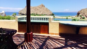 A beachside patio with a wooden table and chairs, a thatched roof, and a view of the sea and distant hills.