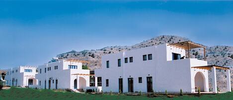 A row of white buildings with flat roofs, a wooden pergola on the right, and a mountainous backdrop.