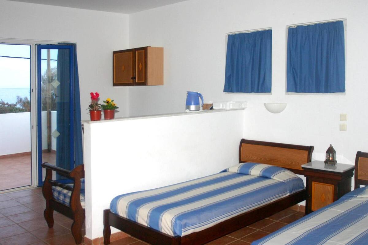 Un dormitorio con cama, una mesita de noche de madera, una pequeña mesa con lámpara, una ventana con cortinas azules, una puerta que da a un balcón y una zona de kitchenette con hervidor y cafetera.