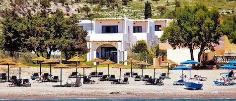 A coastal hotel with a white building, beach umbrellas, sun loungers, and a view of the sea.