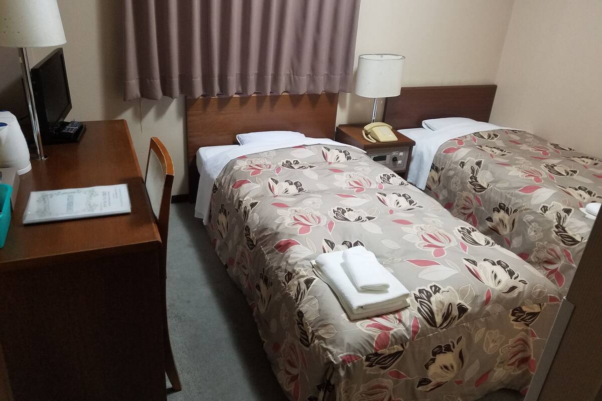 A hotel room with two beds, a desk with a lamp, a telephone, and a window with curtains.