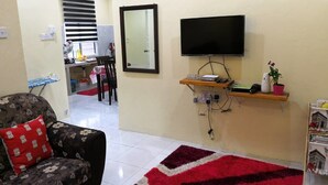 Un soggiorno con un divano, un tappeto rosso, una TV montata a parete, un piccolo ripiano con una pianta e una porta che dà su un'altra stanza.