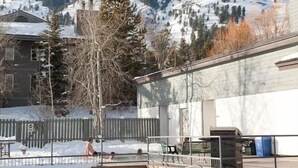 Zona de piscina con nieve en el suelo, rodeada de edificios y montañas al fondo.