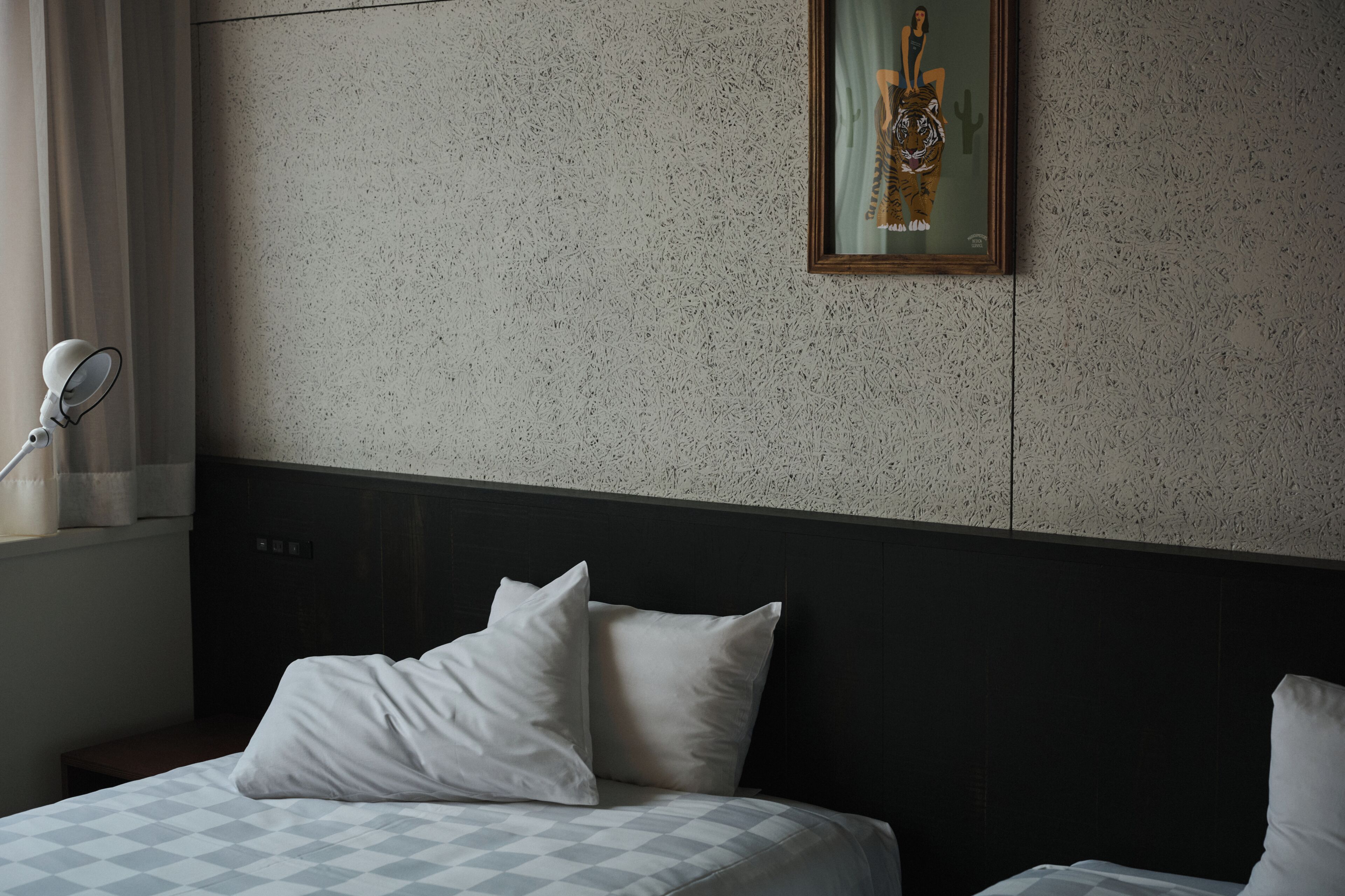 A hotel room with a bed, pillows, a headboard, a wall-mounted picture, and a window with curtains.
