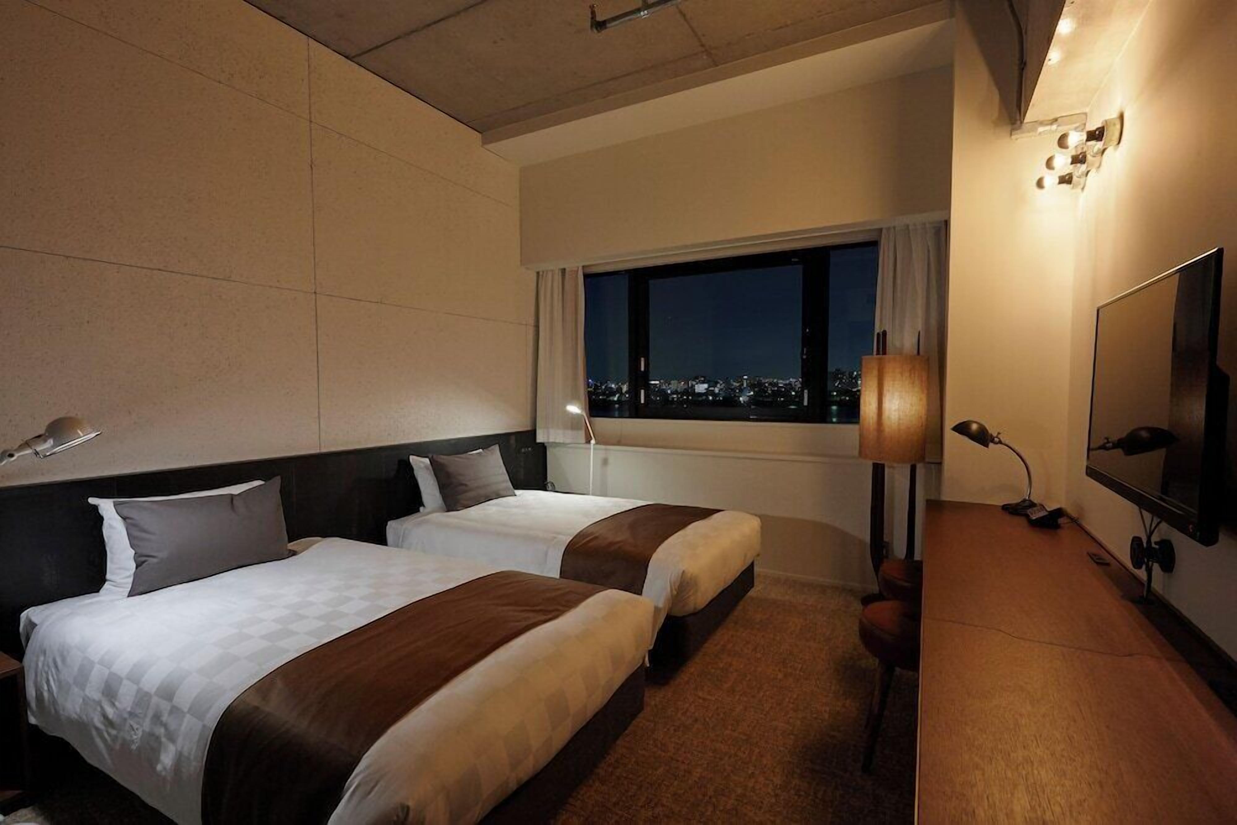 A hotel room with two beds, a large window with a city view, a desk with a lamp, and a TV.