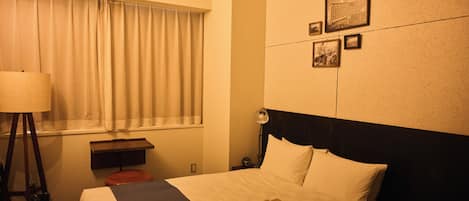 A hotel room with a bed, a bedside table, a lamp, a window with curtains, and a wall with framed pictures.