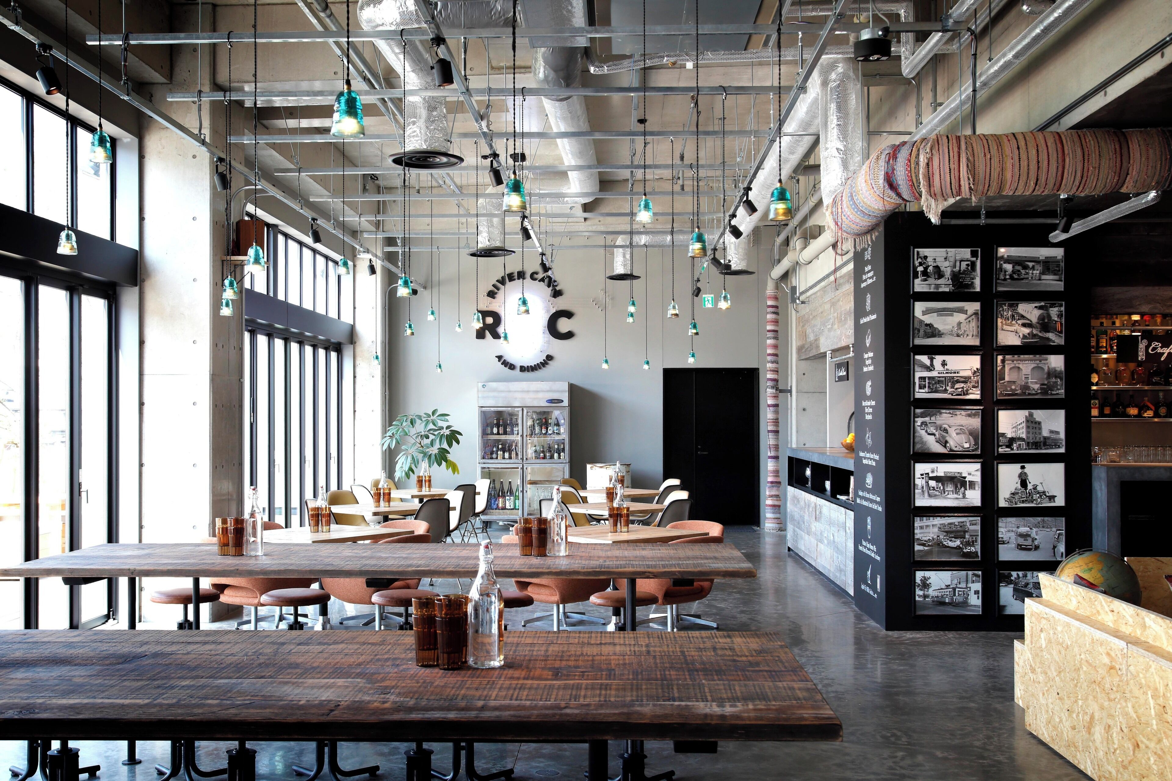 Uma sala de jantar espaçosa com pé direito alto, dutos aparentes e iluminação pendente. Mesas com cadeiras estão dispostas, e uma área de bar é visível à direita.