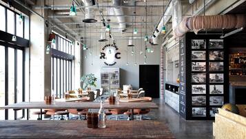Uma sala de jantar espaçosa com pé direito alto, dutos aparentes e iluminação pendente. Mesas com cadeiras estão dispostas, e uma área de bar é visível à direita.