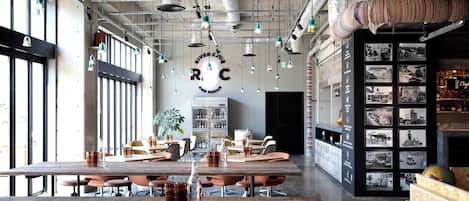 A spacious dining area with high ceilings, exposed ductwork, and pendant lighting. Tables are set with chairs, and a bar area is visible on the right.