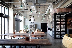 Un comedor amplio con techos altos, conductos expuestos e iluminación colgante. Las mesas están dispuestas con sillas, y se observa una barra a la derecha.