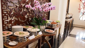 A buffet setup with various dishes labeled, a decorative wall, and a potted orchid plant. - Bazan Xanh Hotel (Buon Ma Thuot)