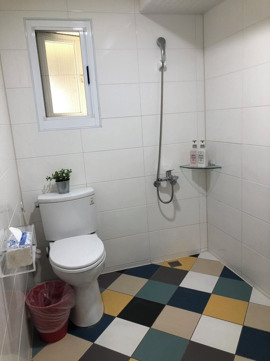 A bathroom with a white toilet, a shower, a small shelf with toiletries, and a checkered floor.