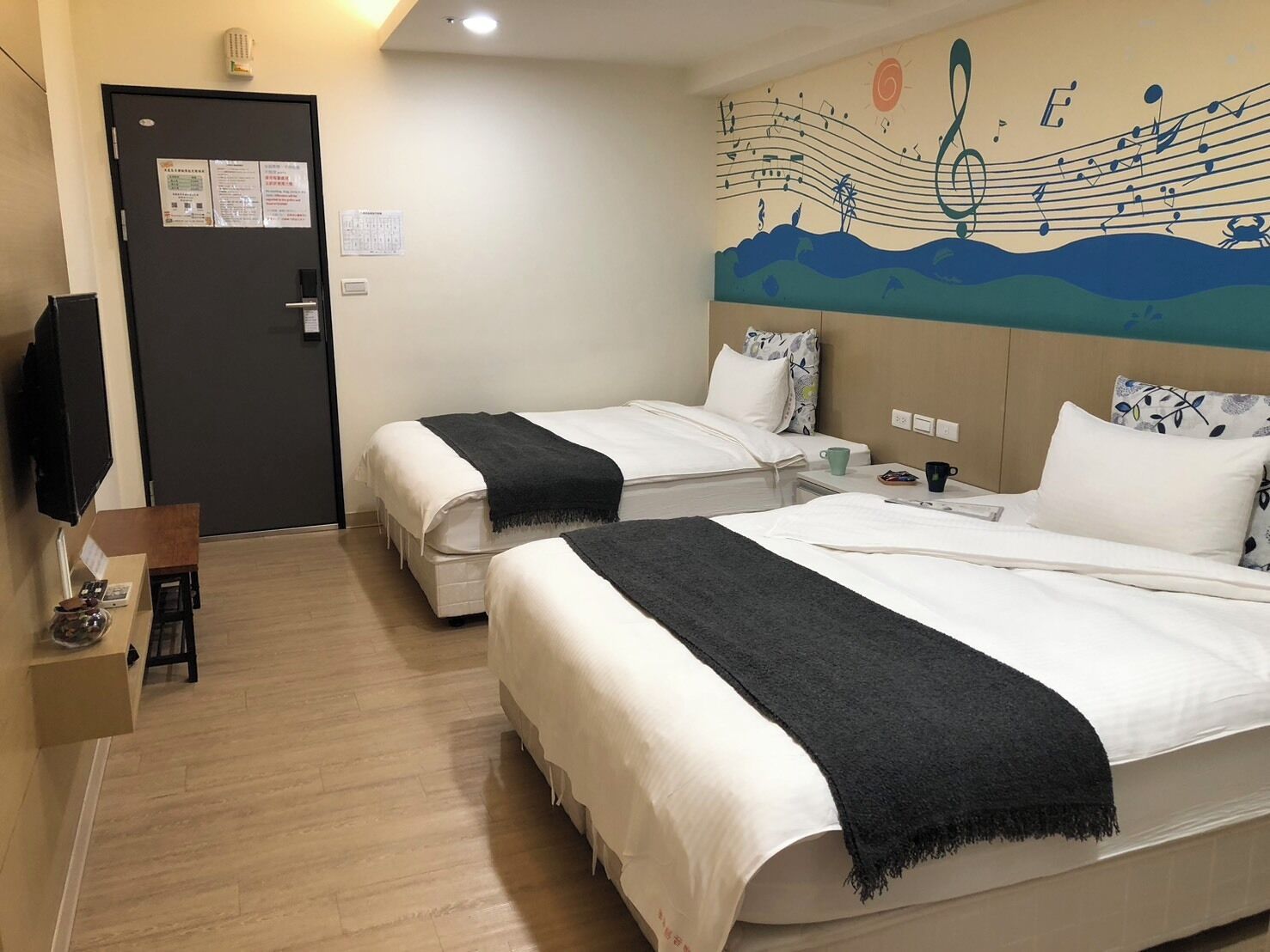Quarto de hotel com duas camas, uma TV de tela plana, piso de madeira e um mural na parede.