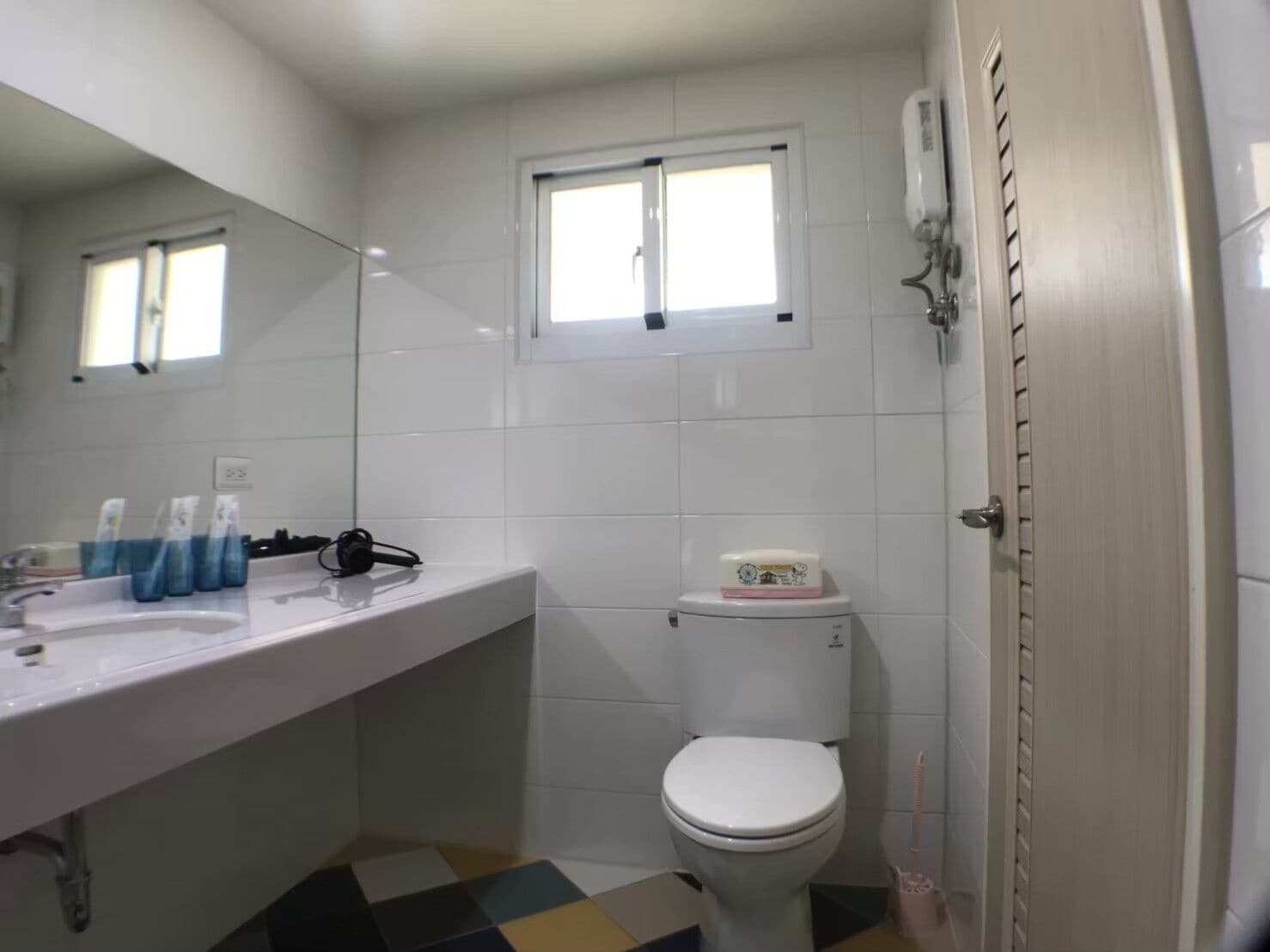 Et badeværelse med en hvid vask, toilet og spejl. Der er et vindue over toilettet, og en brusehoved på væggen.