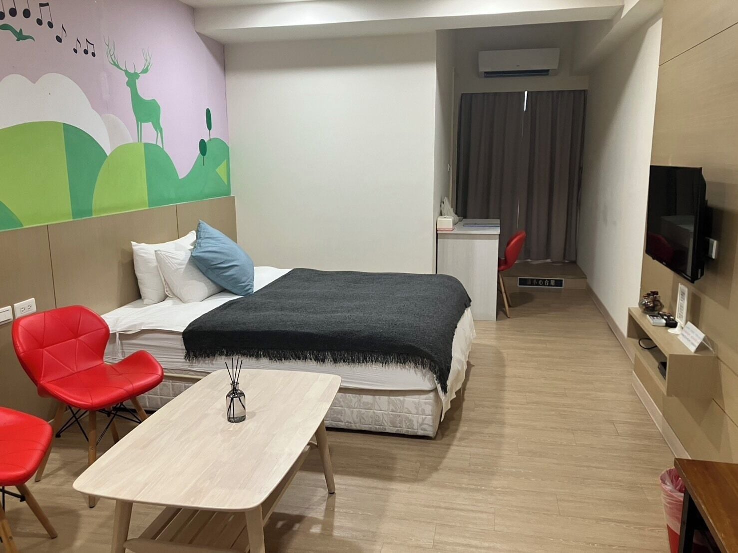 A modern hotel room with a bed, a desk, a TV, and red chairs.