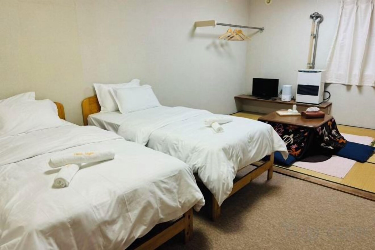 A hotel room with two beds, a desk, a chair, a lamp, and a small shelf with a laptop and a cup.