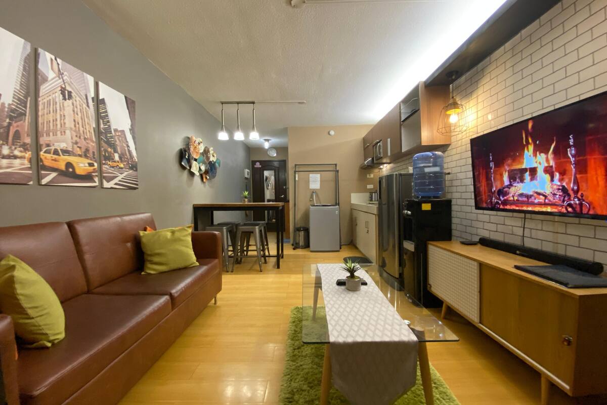 Uma sala de estar com sofá de couro marrom, mesa de centro de vidro e televisão exibindo uma cena de lareira.