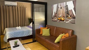 Habitación de hotel con una cama, un sofá de cuero marrón, una mesita con una planta y una pared con una fotografía de un paisaje urbano.