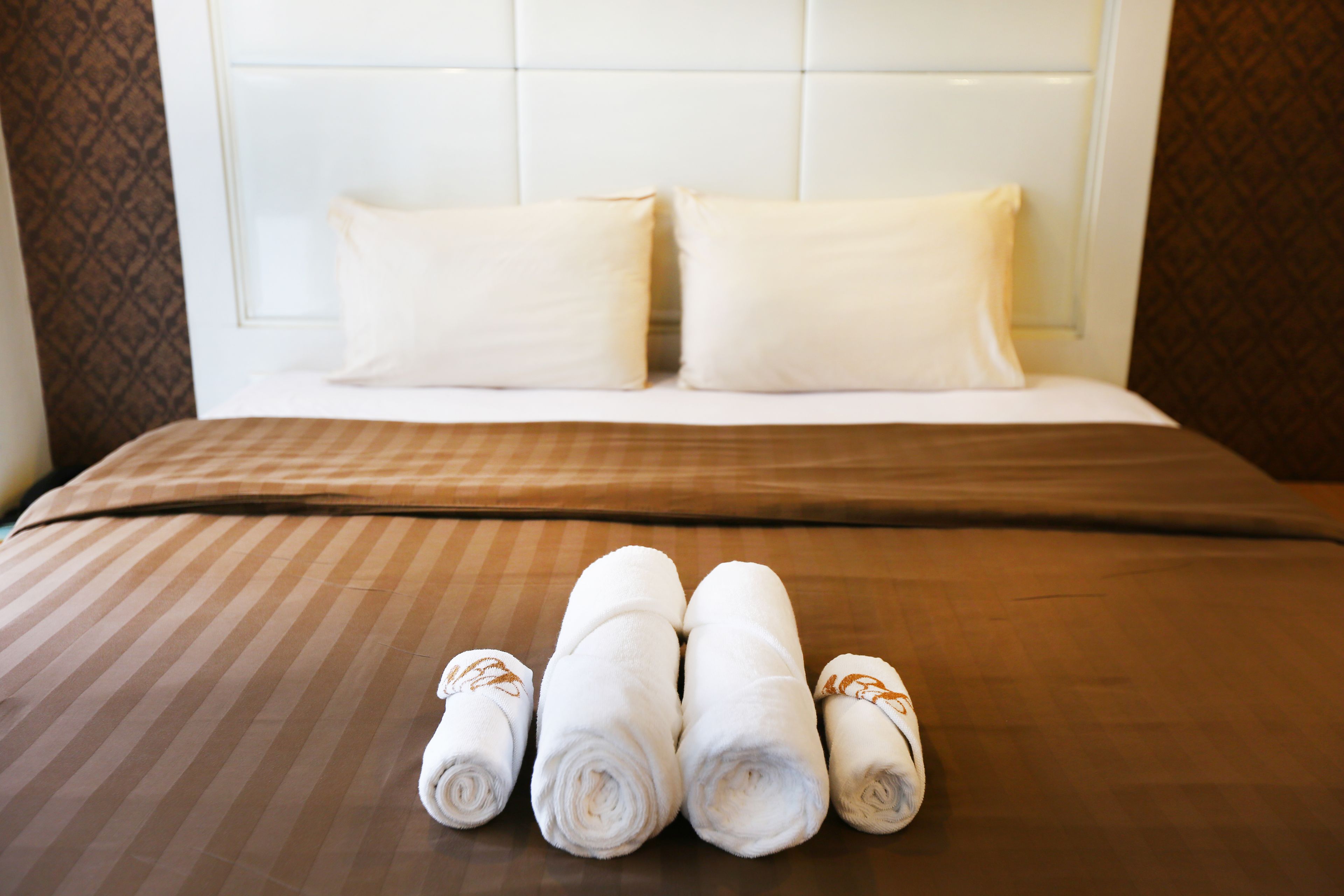 A neatly made bed with a brown striped bedspread and two white pillows. In front of the bed, there are four rolled white towels with brown leaf patterns.