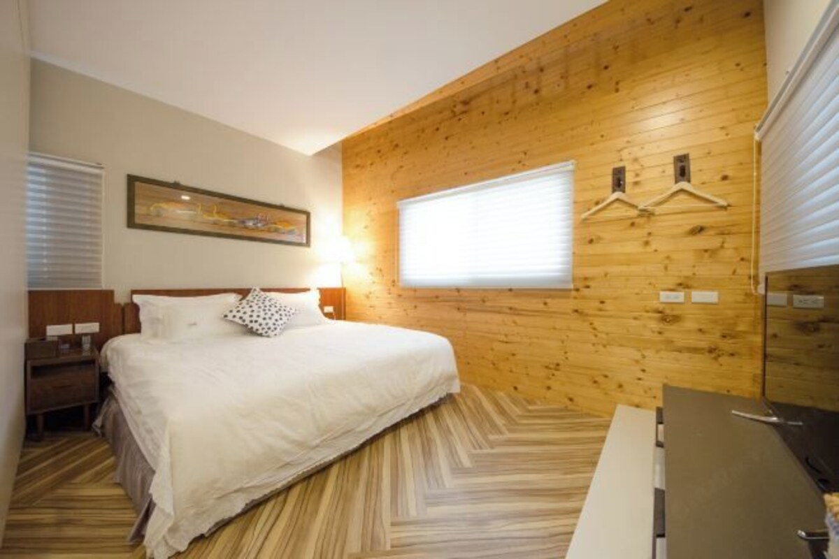 A bedroom with a wooden wall panel, a bed, a nightstand, a bedside table, and a window with blinds.