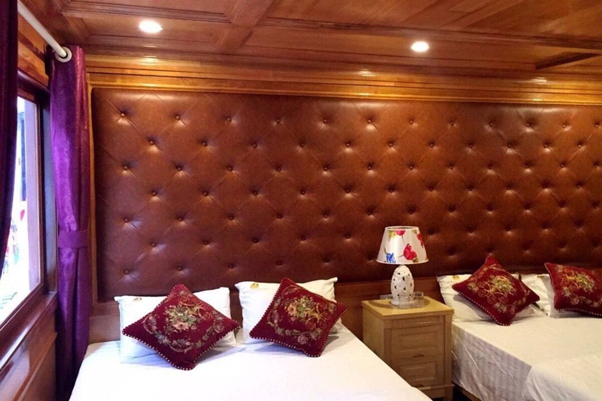 Une chambre d’hôtel avec une tête de lit capitonnée, deux lits avec une literie rouge et blanche, une table de chevet avec une lampe et une fenêtre ornée de rideaux violets.