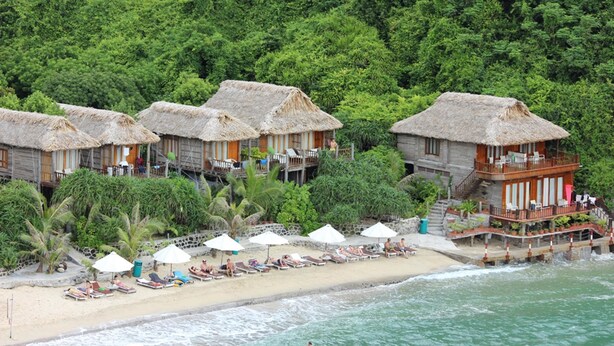 A beachfront hotel with thatched-roof bungalows, a sandy beach, and a lush green forest backdrop. - Cat Ba Sisters Legend Cruise (Hai Phong)
