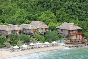 A beachfront hotel with thatched-roof bungalows, a sandy beach, and a lush green forest backdrop. - Cat Ba Sisters Legend Cruise (Hai Phong)