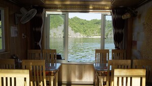 A boat interior with wooden chairs, a table, a fan, and a view of a lake with a boat in the distance. - Cat Ba Sisters Legend Cruise (Hai Phong)