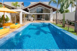 Una casa moderna de dos pisos con un amplio ventanal, piscina y un patio con cojines amarillos.