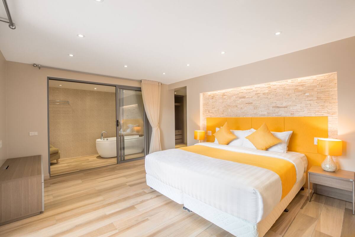 A modern hotel room with a large bed, a glass shower enclosure, and a wooden floor.