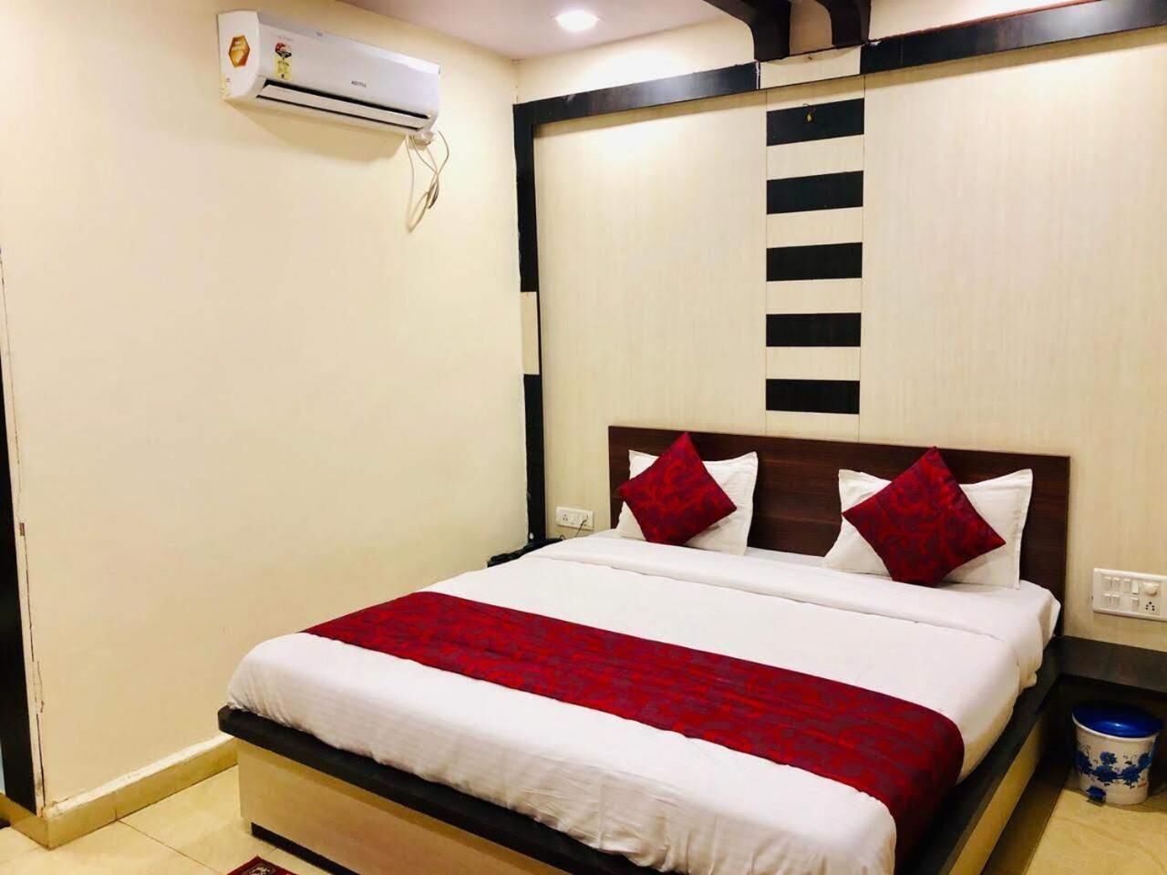 Habitación de hotel con cama, colcha roja y blanca, almohada blanca, almohada roja, cabecero de madera, aire acondicionado de pared y un cesto de basura pequeño.