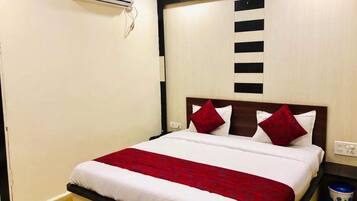 Habitación de hotel con cama, colcha roja y blanca, almohada blanca, almohada roja, cabecero de madera, aire acondicionado de pared y un cesto de basura pequeño.