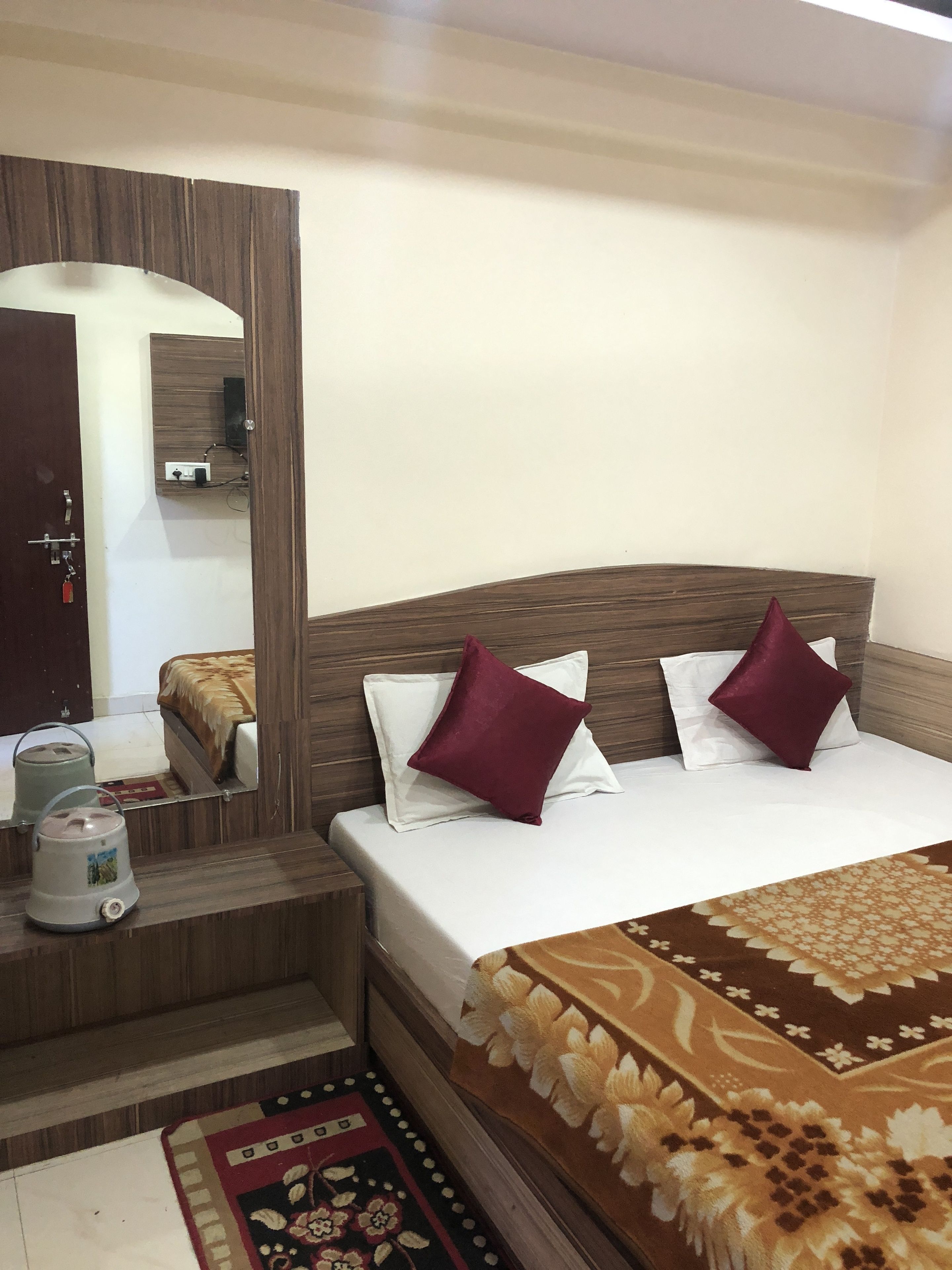 A neatly arranged bedroom with a wooden headboard, a bed with white and maroon pillows, a bedside table with a kettle, and a patterned rug on the floor.